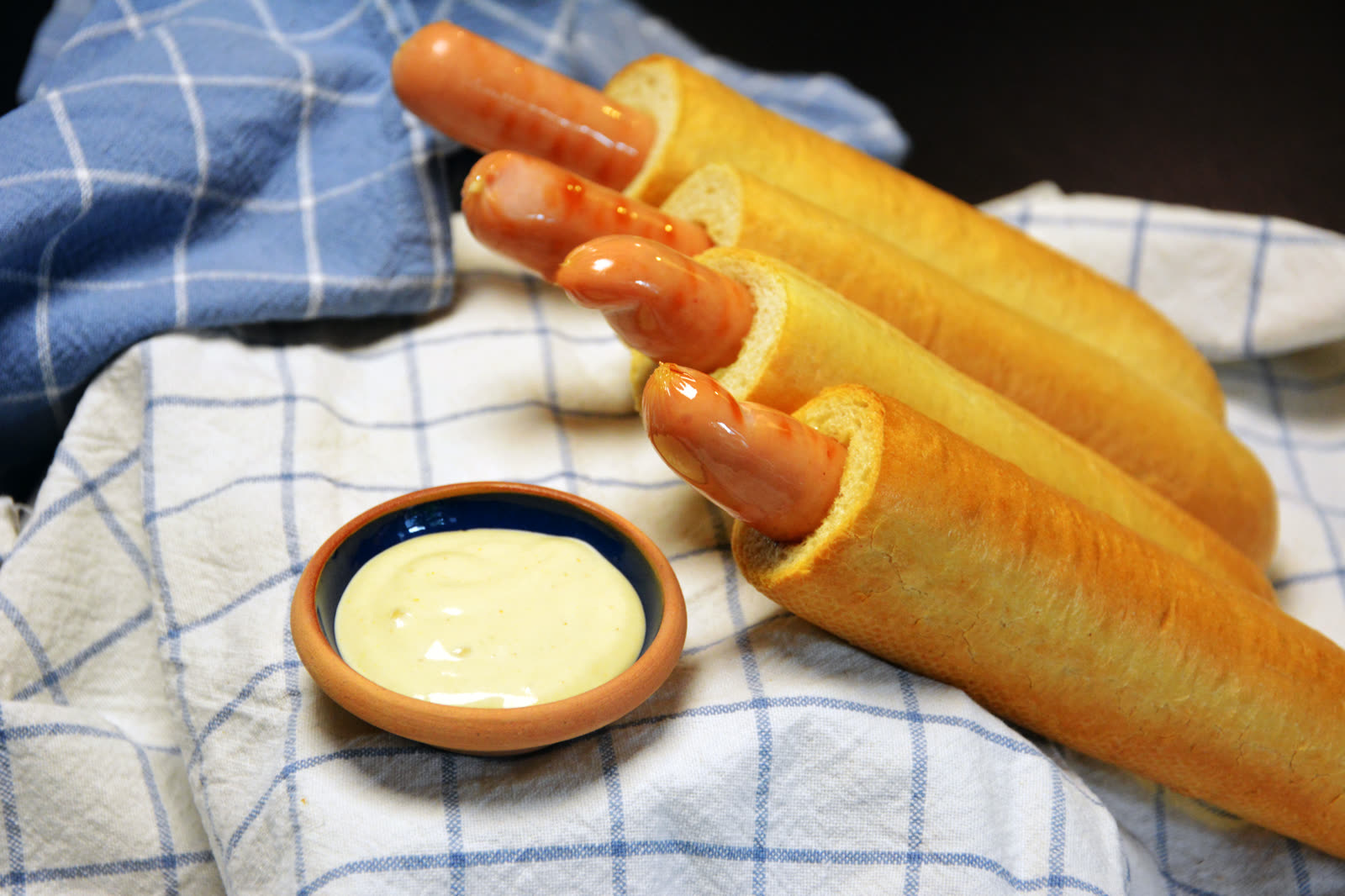 Fyra färdiga franska hotdogs med korven nere i hålet ligger på en blårutig kökshandduk bredvid en liten skål med ljus dressing