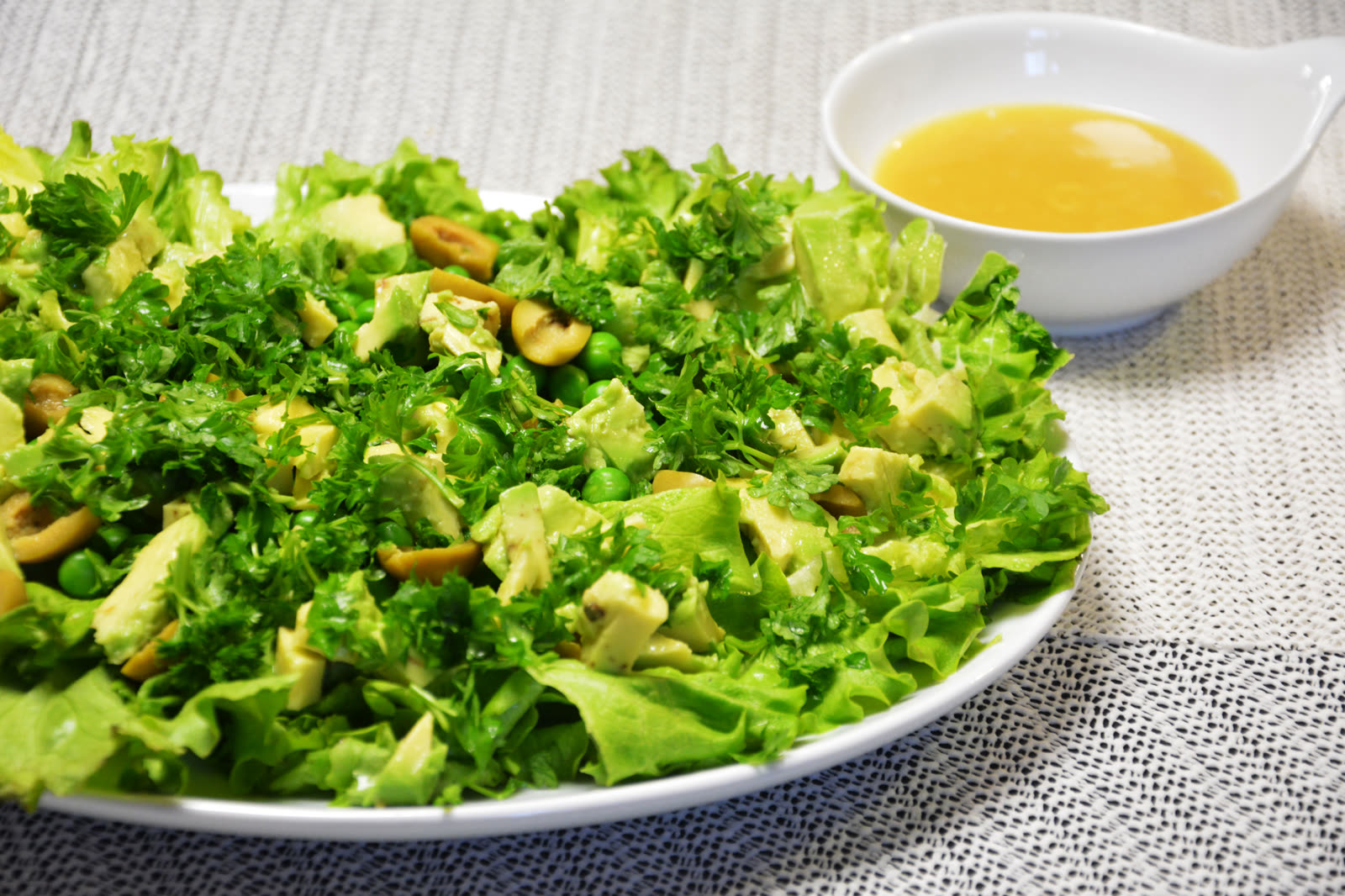 Grönsallad med krussallad, avokado, gröna oliver, ärtor och persilja på ett vitt fat, med vinägretten i en liten skål vid sidan om