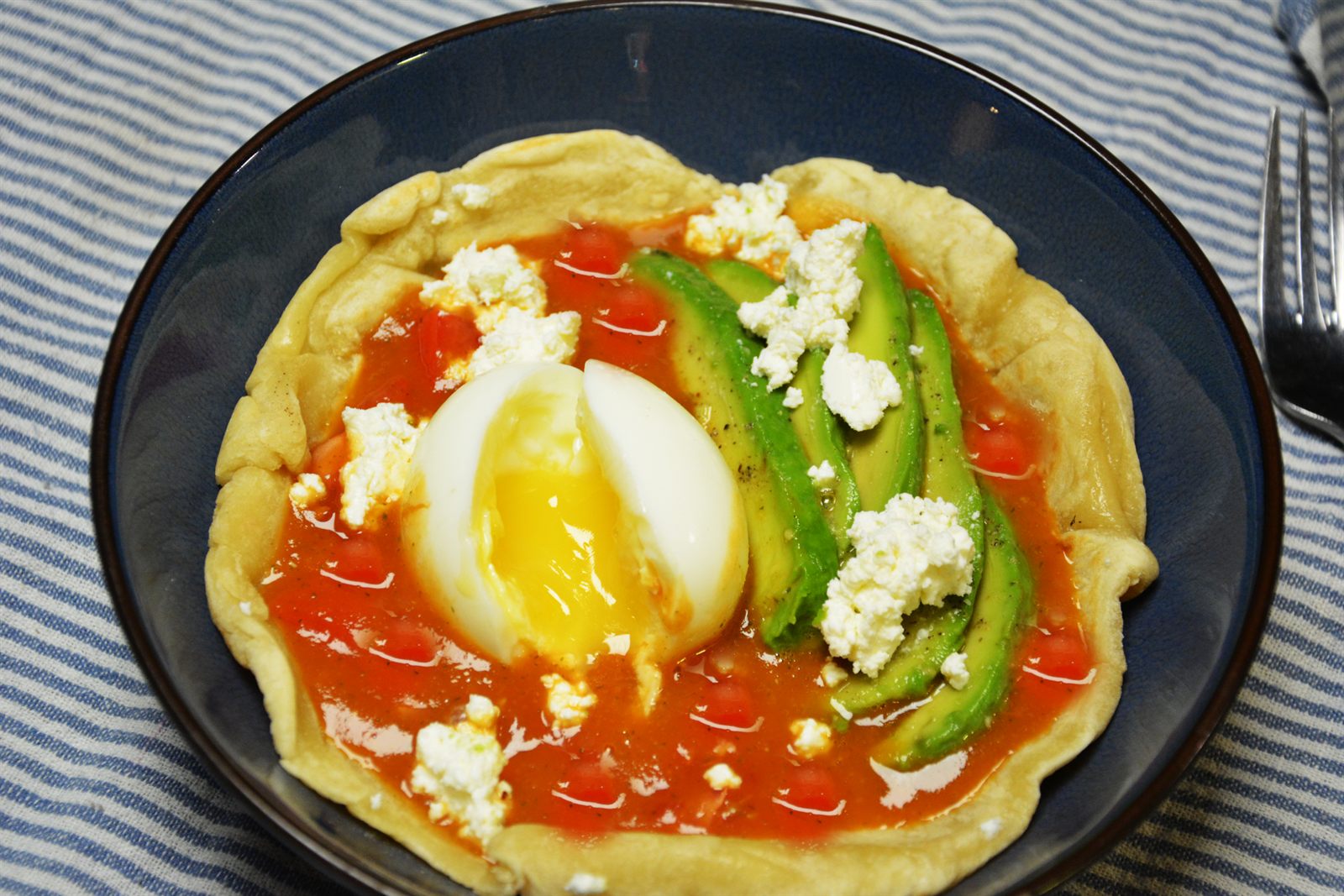 Huevos rancheros i en djup blå skål: en mjuk vetetortilla med salsa roja, ett halverat löskokt ägg, avokadoskivor och smulad fetaost