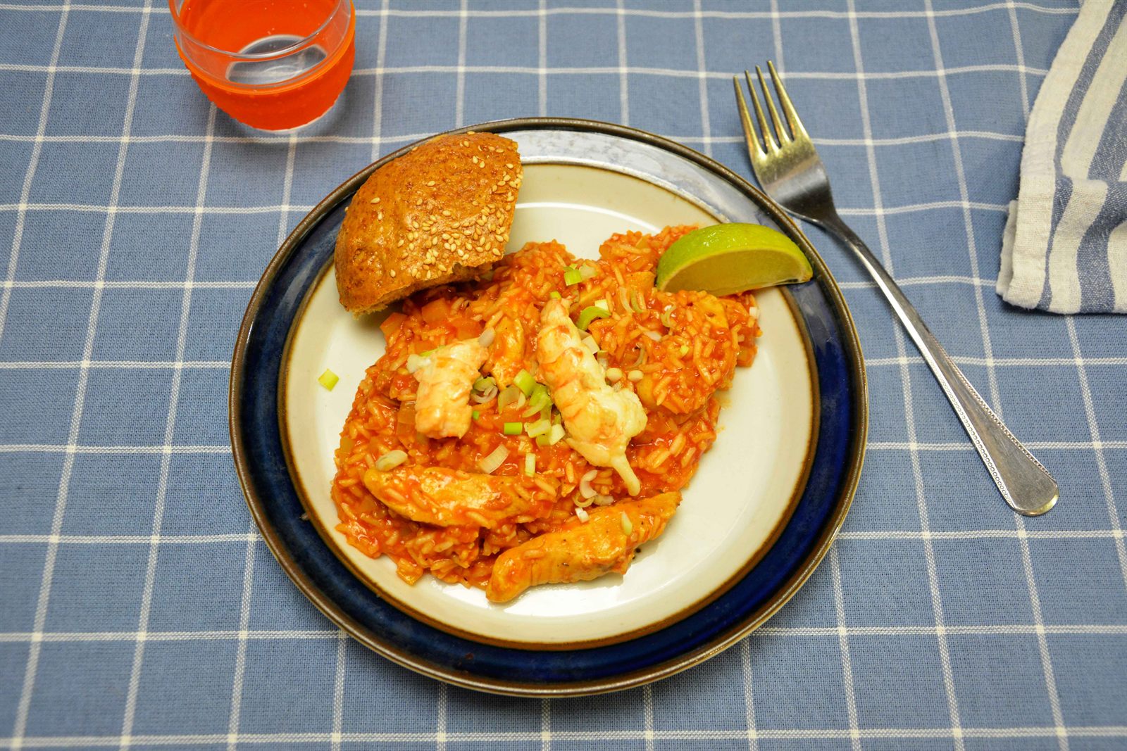Jambalaya med röd tomatris, kycklingstrimlor och havskräftstjärtar upplagd på en vit tallrik med en limeklyfta, hackad salladslök och ett sesambröd