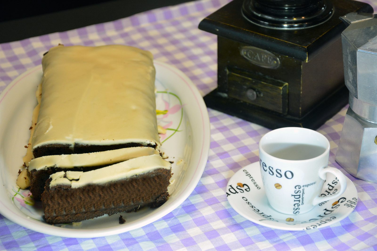 Kaffekaka med ljus kaffeglasyr, skuren i skivor på ett vitt fat, med espressokopp, gammal kaffekvarn och mockakanna bredvid