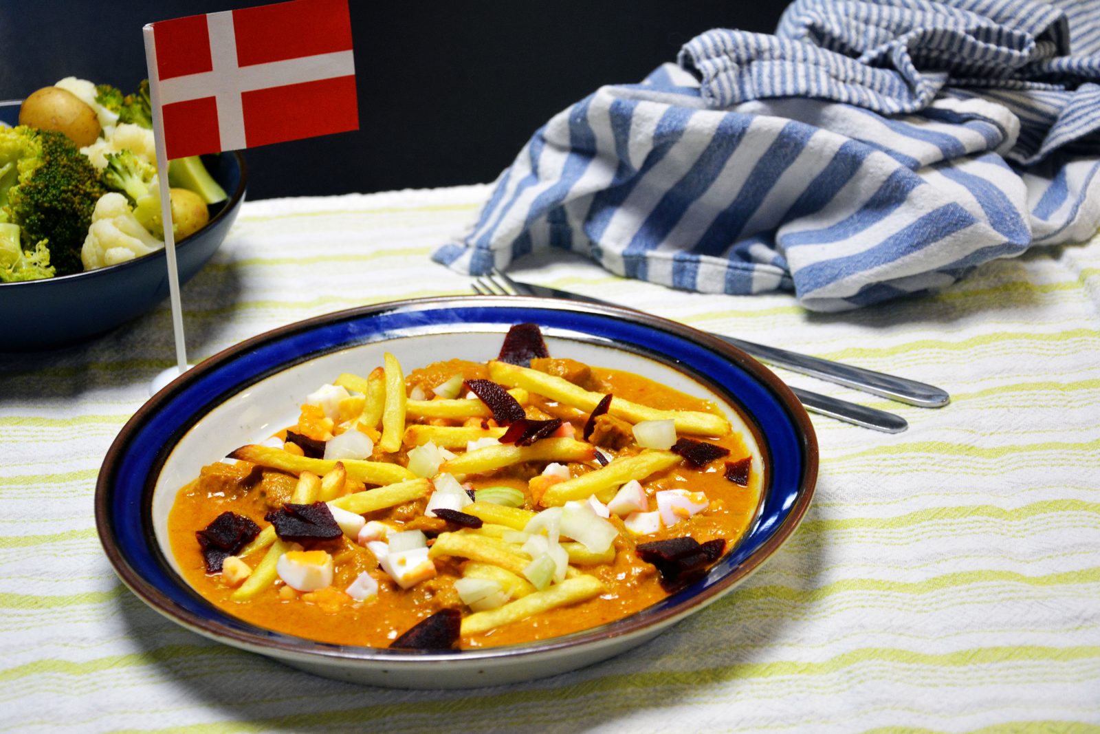 Blåkantad djup tallrik med bitar av fläskfilé i orange gräddsås, toppad med pommes frites, hackat hårdkokt ägg, hackad rå lök och bitar av inlagda rödbetor; till vänster en liten dansk flagga och en mörk skål med små kokta potatisar, broccoli och blomkål, gaffel och kniv, en blårandig kökshandduk och en vit- och gulrandig duk