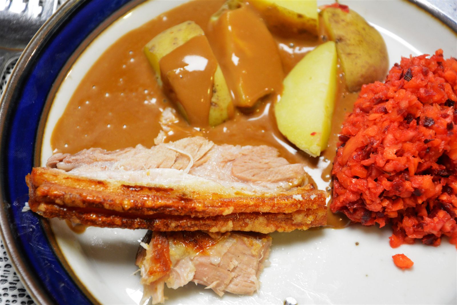 Två skivor långtidsstekt fläsksida med gyllene, knaprig och skiktad svål och ljust kött på en vit tallrik med mörkblå kant; bakom ligger kokt potatis i halvor och fjärdedelar med skal, överhälld med ljusbrun sås, och till höger en hög finhackad råkost i rött och orange.