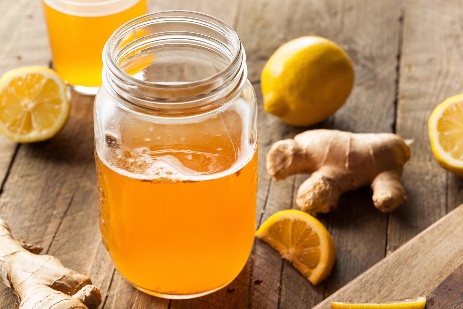 En öppen glasburk utan lock med gyllene, bärnstensfärgad kombucha och lite skum överst, på ett rustikt träbord. Runt burken ligger färsk ingefärsrot, hela och halva citroner och en citronklyfta; i bakgrunden står ett glas med kombucha.