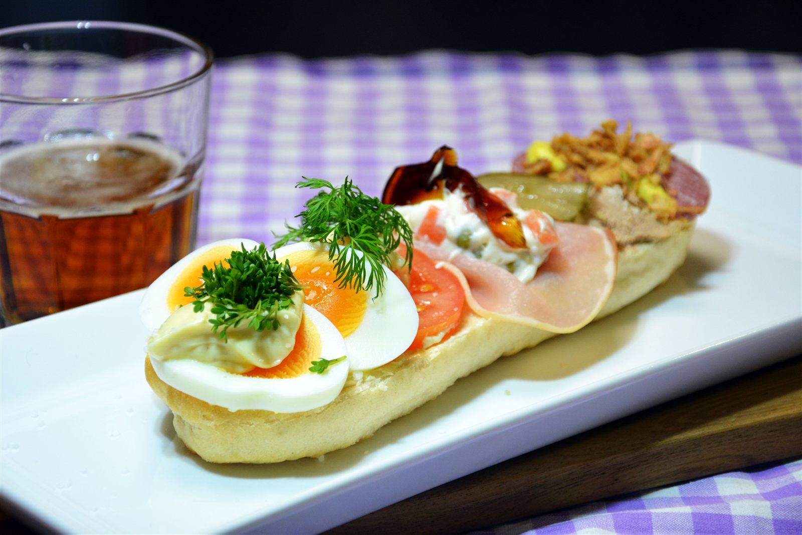 En halv baguette ligger öppen på ett långt, rektangulärt vitt fat. Längst fram ligger kokta äggklyftor med en klick majonnäs och krasse, sedan tomatskivor med en dillkvist, därefter en skiva kokt rökt fläskkarré med italiensk sallad av ärter och morot och en bit sky, och längst bak leverpastej och salami med rostad lök och en skiva saltgurka. Till vänster står ett glas öl. Fatet står på en lila-vitrutig duk mot en mörk bakgrund.