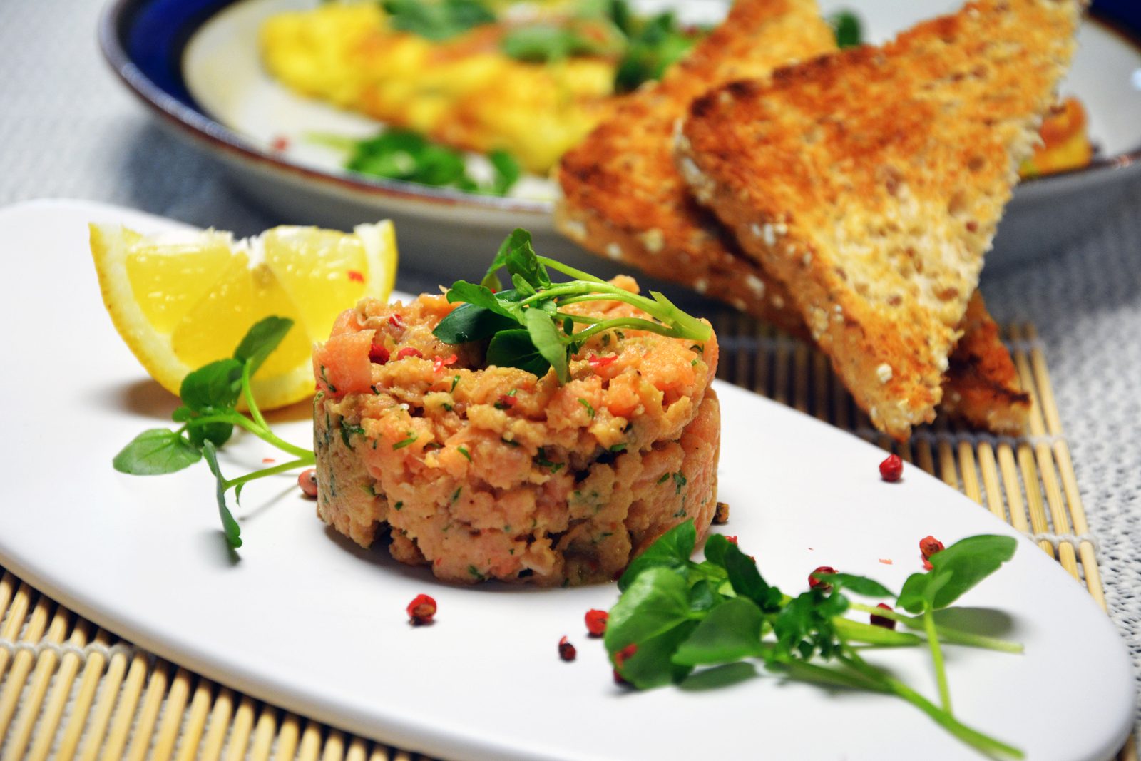 Ett runt torn av laxtartar med dill och rosa pepparkorn, toppat med vattenkrasse, på en avlång vit tallrik med en citronklyfta och lösa rosa pepparkorn; bakom ligger trianglar av rostat kärnbröd, och där står också en tallrik med omelett; den avlånga tallriken står på en bambumatta.